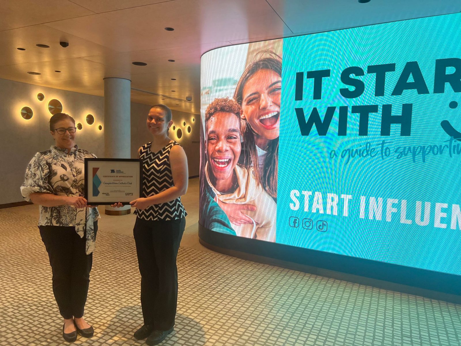Photo of Youth Solutions CEO Geraldine Dean presenting a certificate of appreciation to Campbelltown Catholic Club Marketing Coordinator Jess Satara in front of the campaign digital banner in the Club foyer.